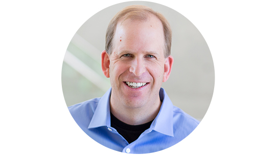 중간색 배경에 옷깃이 달린 파란색 셔츠를 입은 채 웃고 있는 Brian Cross의 사진