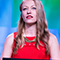 A person wearing a bright red dress speaking to an unseen conference crowd with a soft-focus blue and green background